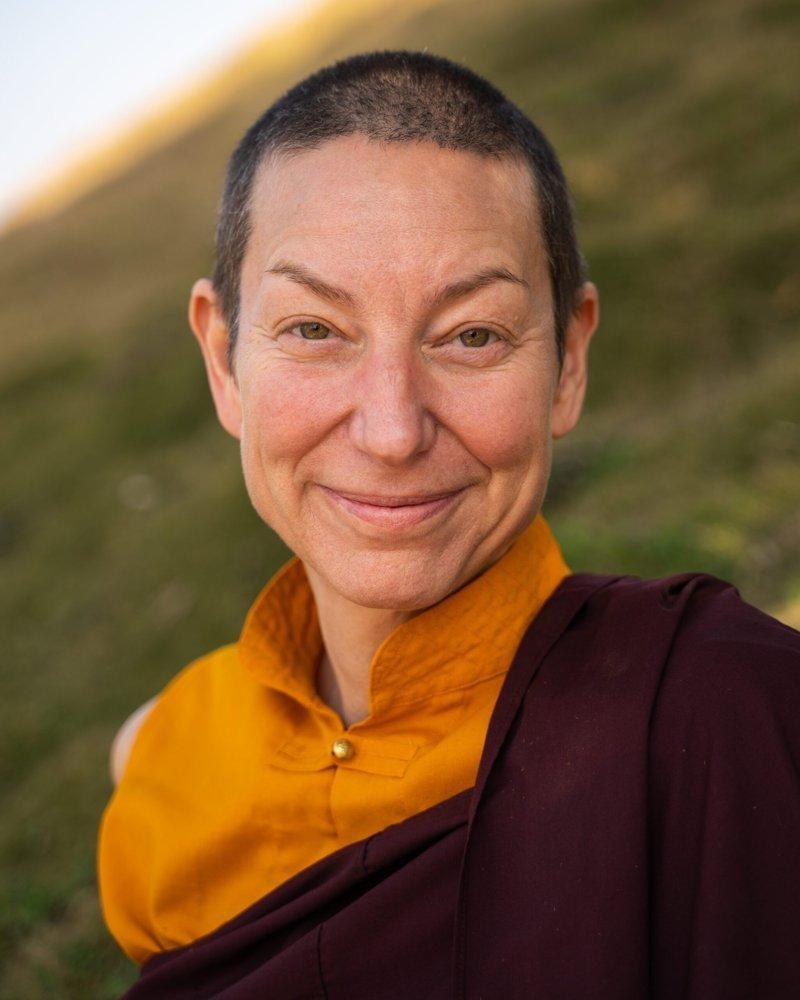 Tsunma Kunsang Palmo smiling, in maroon and gold robes, on a grassy hillside